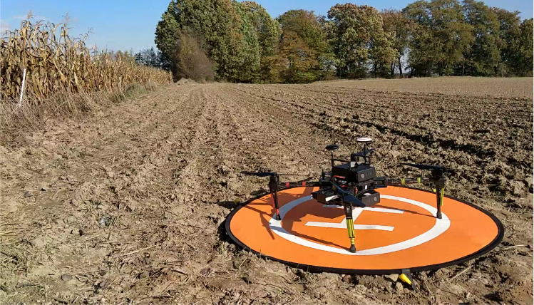 Na polu znajduje się pomarańczowa mata z lądowiskiem dla drona. Dron stoi na macie. W tle widać drzewa.