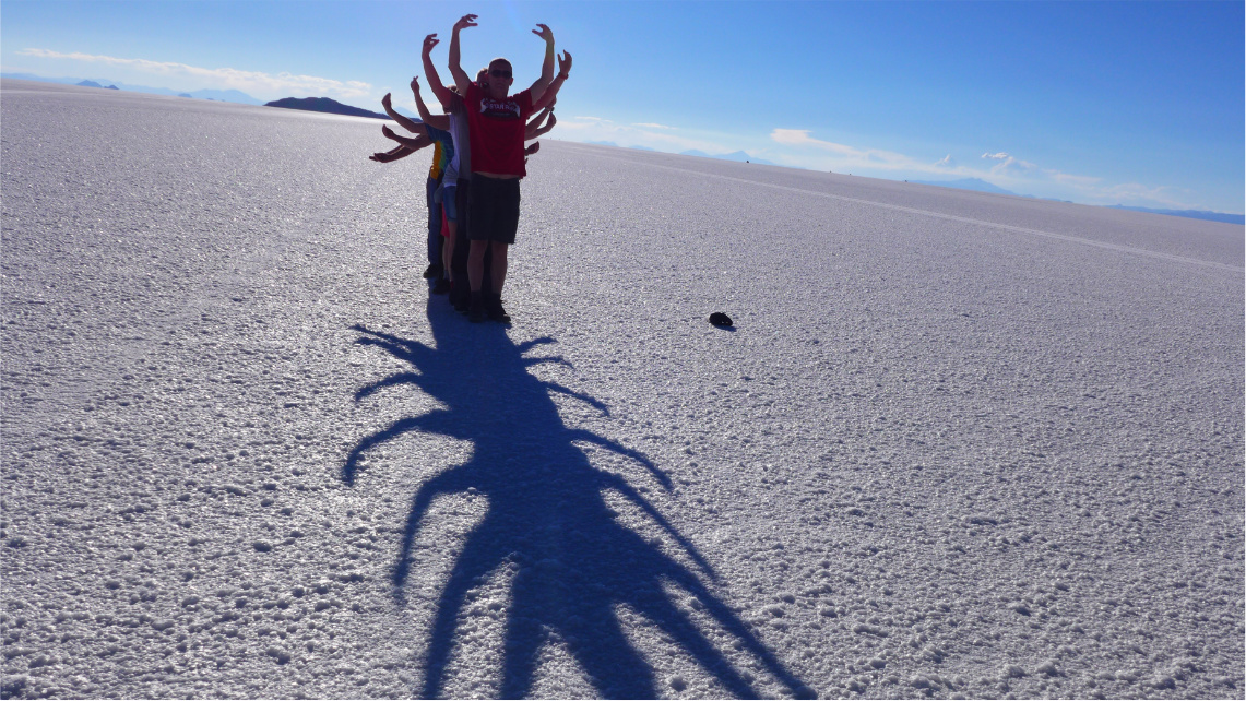 Zdjęcie zrobione pod słońce. Kilka osób stoi jedna za drugą z uniesionymi rękoma, które tworzą cień w kształcie robaka z odnóżami.