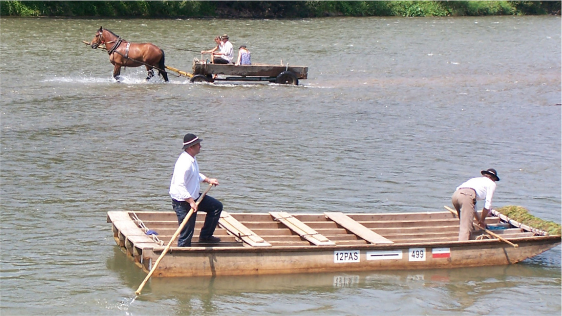 Rzeką płynie tratwa z dwoma mężczyznami na pokładzie. W tle widać zaprzęg konny, który porusza się w wodzie w przeciwnym kierunku niż tratwa.
