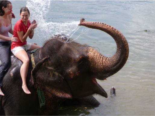 Dwie kobiety siedzą na grzbiecie słonia, który jest zanurzony w wodzie. Słoń oblewa kobiety wodą z trąby.