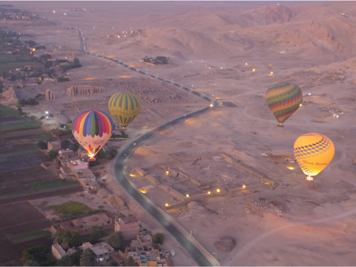 Widok z góry na Dolinę Królów w Egipcie. Na pierwszym planie widać cztery lecące załogowe balony, w oddali znajduje się droga i zabudowania.