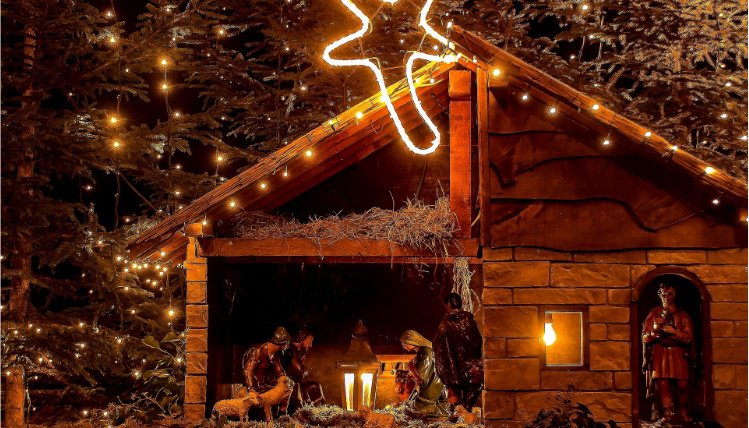 Zdjęcie przedstawiające szopkę bożonarodzeniową z figurkami świętej rodziny. Obok jest drzewko z lampkami. Zdjęcie jest przyciemnione i nastrojowe.
